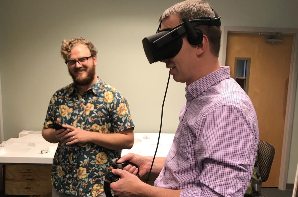 Professor Brad Collett uses virtual reality goggles as a smiling students looks on. 