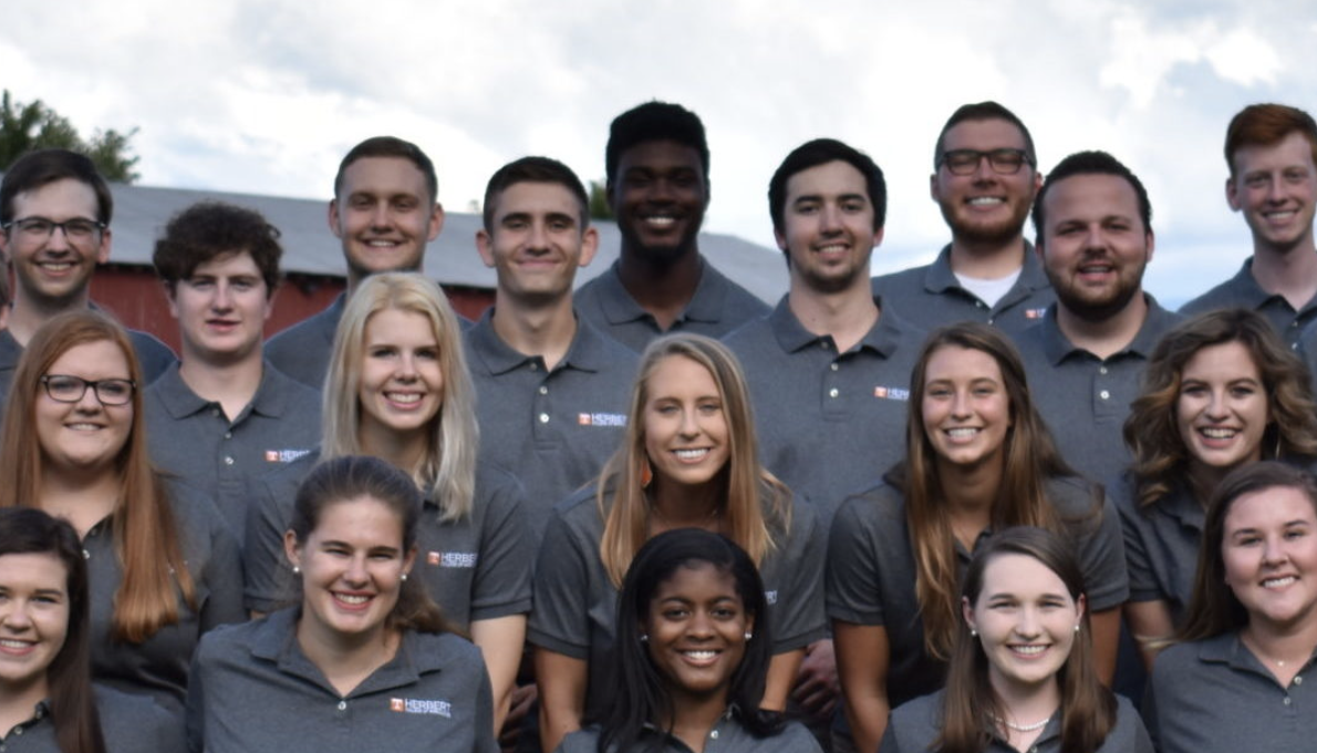The Herbert Ambassadors, a group of diverse students who lead campus activities, pose in matching grey polos in front of a barn