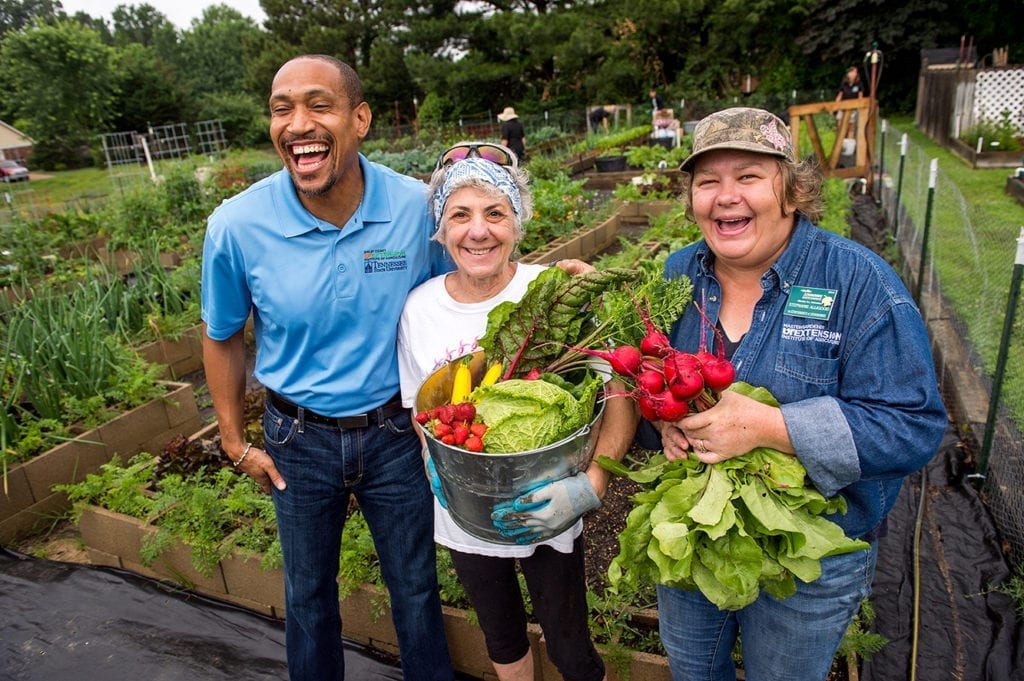 A man and two women stand in front of green rows of garden vegetables, one woman holding a bucket full of produce