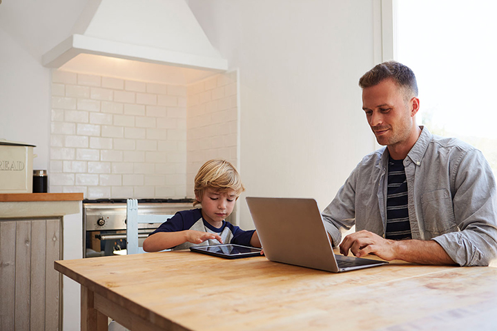 A father works on his laptop at a table while his son sits beside him using a tablet